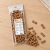 Flat-lay SKU 204603 (Akinu Kosti sušenka se zinkem 1 000 g) — sáček shora na světlém dubovém stole s rozloženými sušenkami-kostičkami. Maketa v Heliosu chybí — reference: BaseLinker čelní foto 1be2e52c + packshot se sušenkami 80011e92 (job 6b286998). 2k 1:1, vybrána v2 ze 2.