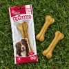 Flat-lay shora na zelené trávě: sáček žvýkadla 204045 (kost z buvolí kůže 16 cm) leží naplocho, vedle dvě volné lisované kosti. Přirozené denní světlo, fotorealistické.