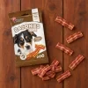 Flat-lay SKU 204731 (Akinu BACONIES Slaninky 85 g) — sáček a slaninky shora na dřevěných prknech jako na BaseLinker lifestyle fotkách (63b5497d), rozložení dle flat-lay 173011. Obal z BaseLinker packshotu 8453dd67 (maketa v Heliosu chybí). 2k 1:1, v1 ze 2.