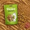 Flat-lay Akinu PAPKY Kostička žlutá 100 g (174027) — pohled shora na zralé obilí, sáček + pamlsky + obilí + malá hrstka syrového masa. Poměr prvků odpovídá složení (obiloviny na 1. místě), maso bez konkrétního druhu.