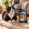 Lifestyle Akinu Masíčka Premium Kachní plátky pro kočky mramorované 50 g (282055) — černobílá domácí kočka v běžném českém obýváku se spokojeně olizuje, vedle ní na zemi leží pár plátků pamlsku a otevřený sáček; teplé odpolední světlo. Varianta b2 ze 2.