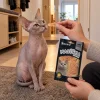 Lifestyle Akinu Masíčka Premium Kuřecí plátky pro kočky mramorované 50 g (282054) — kočka sphynx klidně sedí na koberečku v běžné české předsíni a čeká na pamlsek podávaný z prstů majitele, který drží sáček; teplé podvečerní světlo. Varianta s2 ze 2.